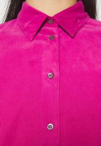 Chemise en velours côtelé fuchsia avec un col classique, cinq boutons ornés de métal argenté, et une texture côtelée verticale.