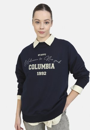 Sudadera de color marino con cuello crema, presenta texto gráfico "Bienvenido a Nueva York" y "COLUMBIA 1992" en colores contrastantes. Corte clásico.
