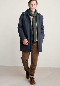 Abrigo de marino impermeable con botones, cierre de cremallera y acentos en los hombros; bufanda de punto verde; camisa de cuadros; pantalones de pana marrones; botas marrones.