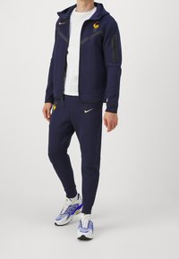 Marinen träningsdräkt tillverkad av mjukt tyg, zip-up jacka med huva och logotyp, i kombination med tapered byxor och mönstrade sneakers.