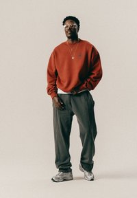 Rød sweatshirt med logo, grå sweatpants med synlige sømme og sølvfarvede løbesko med branding. Briller med klare linser som tilbehør.