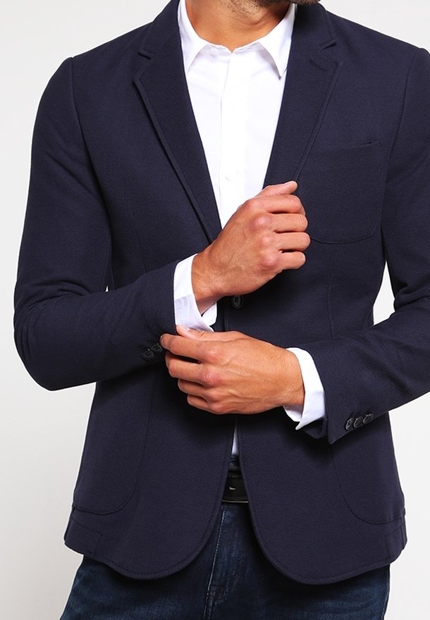 Homme ajustant le poignet d’un blazer bleu marine porté sur une chemise blanche, jean bleu visible à la taille, sur un fond clair uni.