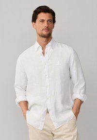Homme aux cheveux bruns portant une chemise blanche à boutons avec les manches retroussées, pantalon beige, debout devant un fond gris clair uni.
