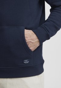 Sudadera azul marino de mezcla de algodón con un bolsillo tipo canguro y puños de canalé. Presenta una pequeña etiqueta de la marca en la parte inferior izquierda.
