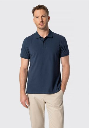 Hombre con cabello corto y rizado que lleva una camisa polo azul marino y pantalones beige, de pie con una mano en el bolsillo contra un fondo liso.