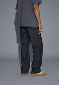 Jeans in denim blu scuro con gambe larghe e cuciture a contrasto, abbinati a una t-shirt grigia e dettagli in grigio e bianco nelle scarpe da ginnastica.