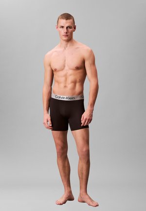 Brązowe bokserki wykonane z gładkiego materiału z szarym logo Calvin Klein na pasku. Model stoi boso na szarym tle.
