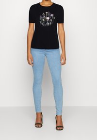 Camiseta negra de manga corta con bordado floral en varios colores en el centro, combinada con jeans ajustados de un azul claro y tacones negros.