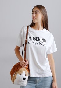 Bijela pamučna majica s natpisom "MO5CH1NO JEANS" otisnutim u crnoj boji. Smeđa plišana torba u obliku psa s ušima i nosom, na smešnom remenu.