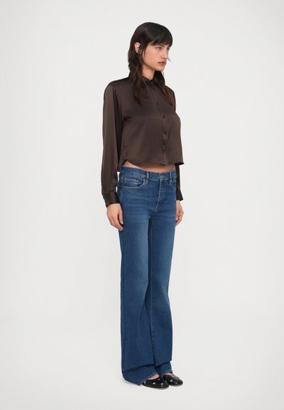 Egy nő áll sötétbarna szatén ingben, magas derekú kék bő szárú farmernadrágban és fekete, ezüst szegecses mokaszinban.