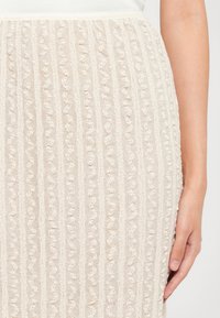 Jupe en tricot beige avec des côtes verticales et des motifs ondulés, fabriquée en tissu texturé, avec une coupe ajustée et une taille élastique.