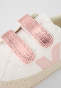 Sapatilha branca com tiras de velcro metálicas cor-de-rosa, parte superior em couro, sola em borracha e detalhe de logótipo rosa na lateral. Design limpo e minimalista.