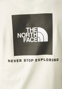 Černý čtverec s bílým logem "The North Face" nad textem "Nikdy nepřestávejte objevovat" na světlem tkaném pozadí.