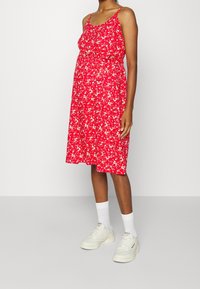 Abito maternità con stampa floreale rossa, spalline sottili, taglio al ginocchio, tessuto fluido e vestibilità rilassata, abbinato a sneakers bianche e calzini.