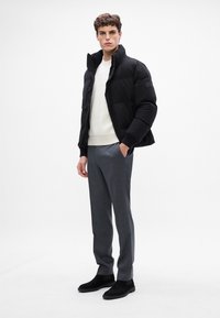Jachetă puffer neagră cu guler înalt, pulover alb cu dungi dedesubt, pantaloni gri croiți și cizme ankle din suede negre.