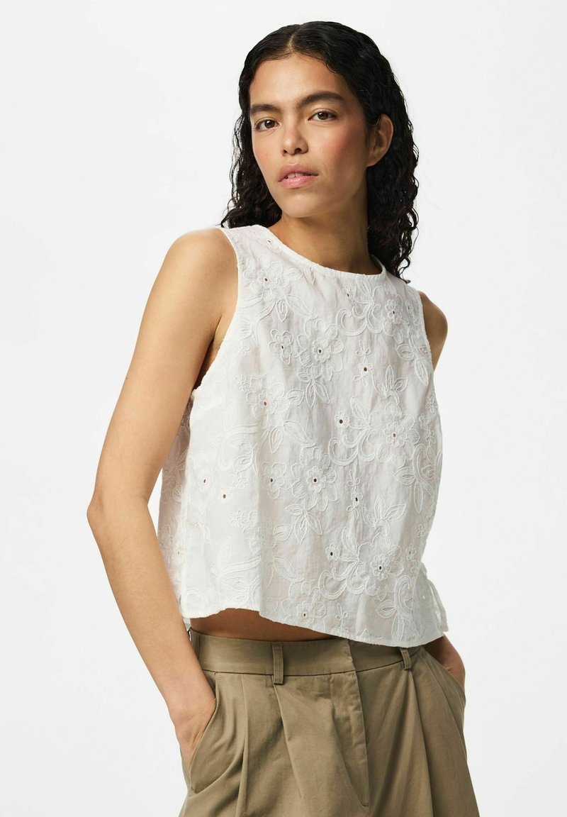 Femme aux cheveux bouclés foncés portant un haut blanc sans manches brodé de fleurs et un pantalon beige taille haute, mains dans les poches, fond neutre.
