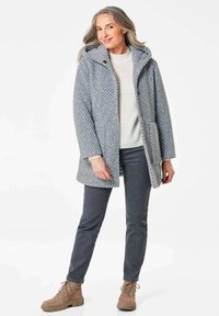 Grauer gestrickter Mantel mit Kapuze, ausgestattet mit einem strukturierten Muster, vorderen Taschen und Knöpfen. Getragen über einem hellen Pullover und dunklen Jeans.