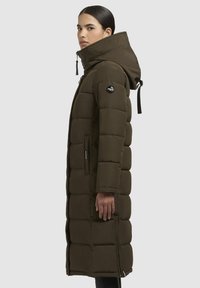 Cappotto puffer verde oliva con cappuccio, caratterizzato da un design trapuntato, zip laterali e una patch rotonda con logo sulla manica sinistra.