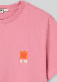 T-shirt rose à manches courtes avec col rond, petit motif de soleil orange, et le mot "SUNSET" imprimé en dessous au centre devant.
