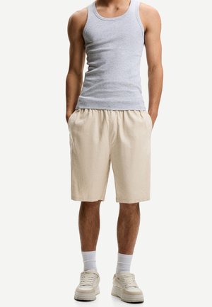 Mann trägt ein graues ärmelloses Hemd, beige Shorts mit Taschen, weiße Socken und beige Sneakers, steht mit den Händen in den Taschen.