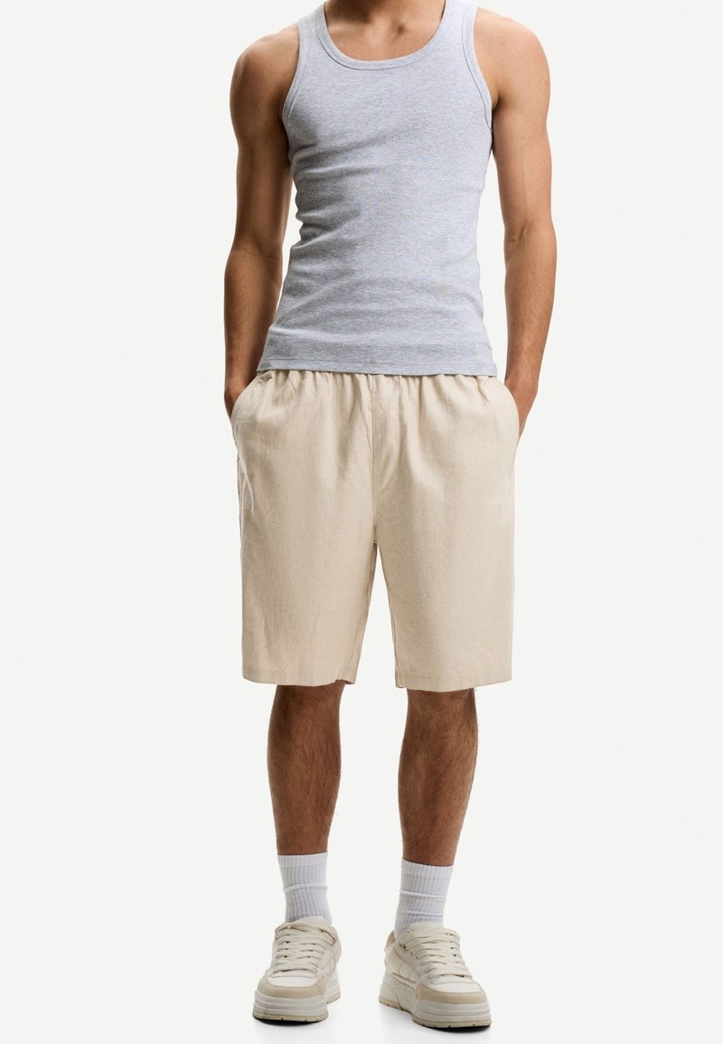 Homme portant un débardeur gris, un short beige avec des poches, des chaussettes blanches et des baskets beiges, debout les mains dans les poches.