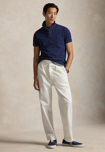 Bărbat în picioare, purtând un tricou polo bleumarin cu mâneci scurte, pantaloni albi cu talie înaltă, adidași albaștri și un ceas de mână pe un fundal bej.