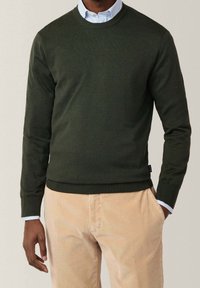 Maglione lavorato a maglia verde scuro con colletto rotondo, polsini e orlo a coste. Indossato sopra una camicia azzurro chiaro, abbinato a pantaloni in velluto a coste beige.