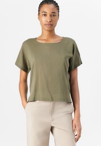 Top verde oliva a maniche corte con scollo quadrato e vestibilità rilassata, realizzato in tessuto leggero, abbinato a pantaloni di colore neutro.