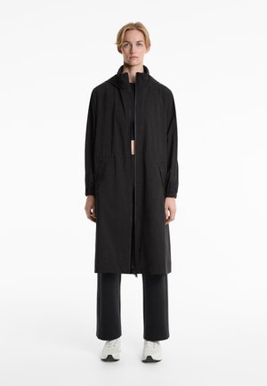Long black lightweight coat with a high collar, elastic cuffs, front zip closure, and side pockets, worn over dark pants and white sneakers.
