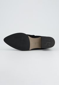 Zapato de mujer de ante negro con punta afilada y suela plana de goma negra. Presenta una plantilla beige con el nombre de la marca grabado en ella.