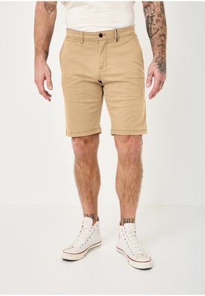 Man wearing beige knee-length shorts and white high-top sneakers, standing with arms relaxed, showing tattoos on arms and legs.