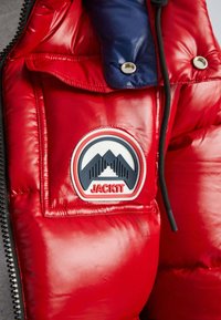 Giacca imbottita rossa con finitura lucida, caratterizzata da un colletto blu, un logo circolare con grafiche montane e dettagli con zip nera.