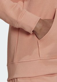 Persikofärgad sweatshirt med en hörnvy som visar det texturerade tyget, ribbade mudd och en framficka. Slät finish med subtila sömdetaljer.