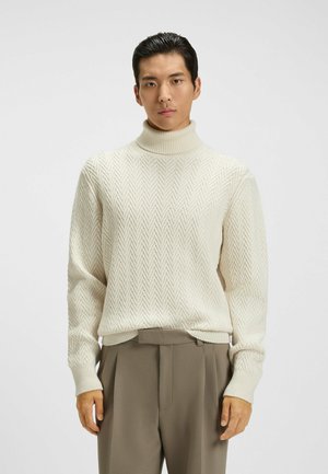 Maglione a collo alto color crema con un motivo herringbone testurizzato, con polsini e orlo ribattuti, abbinato a pantaloni beige a vita alta.