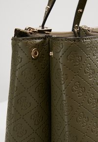Oliven grøn læder håndtaske med præget logo mønster, guldfarvet hardware og dobbelte top hanke. Lynlås lukning øverst.