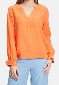 Blusa arancione con texture e scollatura a V, maniche lunghe a sbuffo con polsini arricciati. Indossata con pantaloni azzurri chiari.