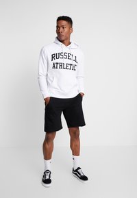 Sudadera blanca con "RUSSELL ATHLETIC" impreso en negro, combinada con pantalones cortos negros, calcetines blancos y zapatillas negras. Diseño simple y atlético.