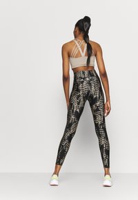 Beige sport-bh med korsade band, kombinerad med svarta leggings med hög midja och ett texturerat mönster i guld och svart, båda använda med vita sneakers.