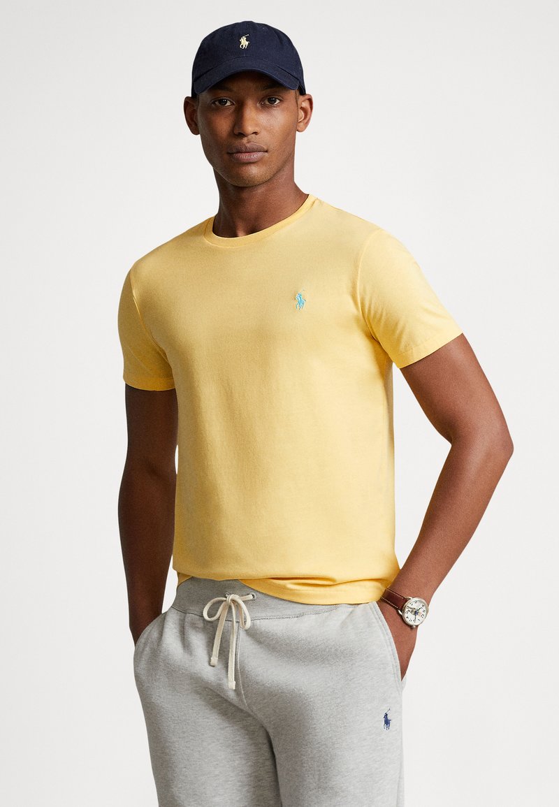 Κίτρινη βαμβακερή μπλούζα με κοντά μανίκια και σ�τρογγυλό λαιμόκοψη, με μικρό τιρκουάζ λογότυπο στο στήθος. Φοριέται με γκρι φόρμα.