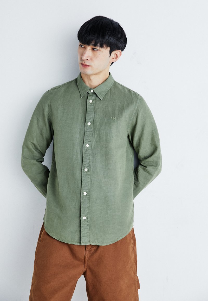 Camicia in lino verde oliva con colletto, ricamo piccolo "Lee" e bottoni bianchi. Pantaloni marroni visibili sotto.