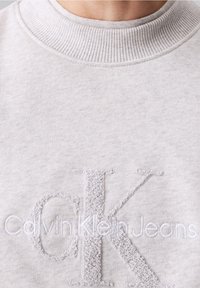 Sudadera gris claro con cuello de canalé. Presenta un "CK" texturizado bordado y "Calvin Klein Jeans" en sutiles puntadas blancas.
