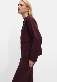 Pulover tricotat de culoare burgundy cu decolteu rotund, mâneci lungi și un buzunar pătrat. Textură netedă, croială lejeră, asortat cu o piesă de îmbrăcăminte de jos similară.