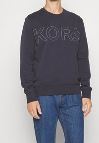 Tmavě modrý svetr z bavlny, s kulatým výstřihem a žebrovanými manžetami. Na přední straně je bílé prošití s logem "KORS".