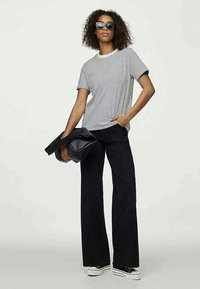 Pruhované bavlněné tričko s krátkými rukávy, černé kalhoty s širokými nohavicemi, černá kabelka a černé sluneční brýle na modelce před jednolitým pozadím.