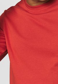 Chemise rouge en coton à manches longues avec un col rond. Texture lisse et forme ajustée visibles, avec des détails côtelés aux poignets et au col.