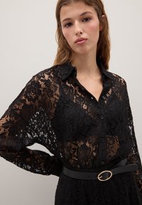 Giovane donna che indossa una camicetta nera in pizzo con bottoni, caratterizzata da un motivo floreale trasparente e una cintura nera con una fibbia ovale dorata.