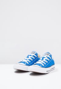 Blå canvas sneakers med vita gummisålar, rund tå och vita snörningar. Har varumärkeslogotyp på tungan och kontrasterande svarta ränder på sålen.