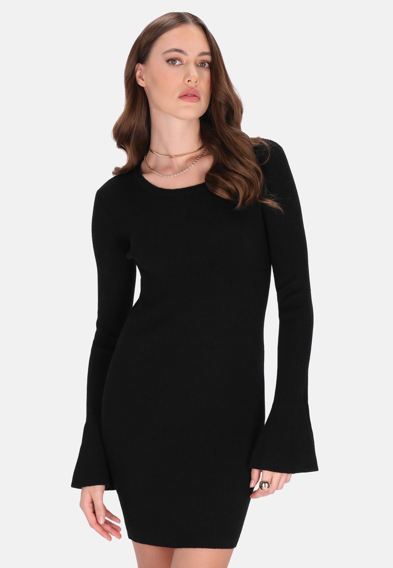 Barna, hullámos hajú nő fekete, hosszú ujjú, testhezálló ruhában, amelyen bővülő mandzsetták vannak, valamint két arany nyaklánc, fehér háttér előtt.