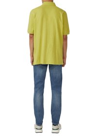 Koszulka polo z krótkim rękawem w jaskrawo limonkowym kolorze, wykonana z teksturowanej tkaniny, w połączeniu z jasnoniebieskimi dżinsami i białymi tenisówkami. Widok z tyłu.