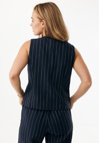 Granatowy top bez rękawów z pionowymi białymi cienkimi paskami, z wysokim dekoltem i dopasowanym fasonem. Gładka faktura tkaniny.